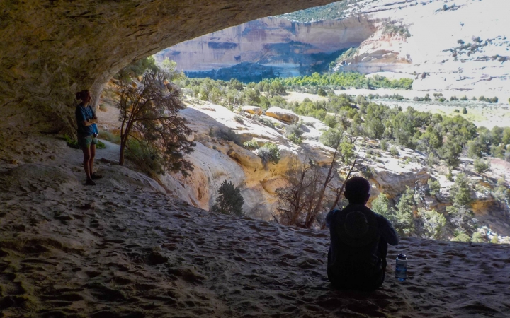 A person rests in the shade of a canyon overlooking a desert area.